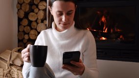 Happy young woman using smart phone surfing social media, checking news sitting near fireplace. Millennial lady drinks hot tea or coffee hold mobile phone, chatting with friends online at wintertime