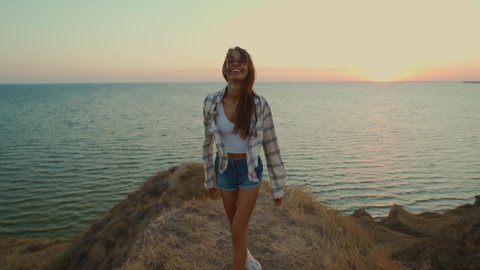 Happy positive emotions cheerful girl laughing and shows kiss on cliff edge with seascape during sunset. Adventure lifestyle, love traveling