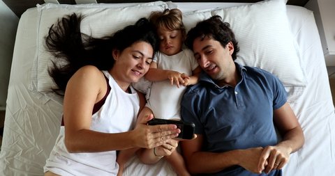 Married millennial couple watching video with infant toddler baby bou in bed
