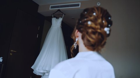 The bride looks at the dress suspended from the ceiling
