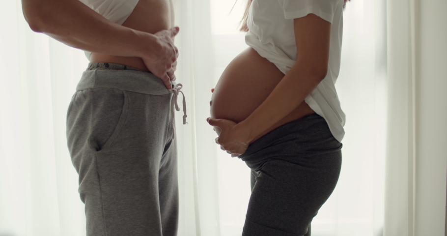 Happy Asian husband and his wife touching her pregnant belly while standing beside a window in their beautiful bedroom.