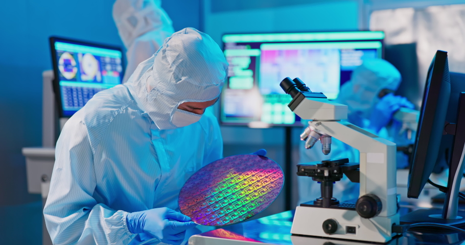 Asian male technician in sterile coverall holds wafer that reflects many different colors with gloves and check it at semiconductor manufacturing plant | Shutterstock HD Video #1083484102