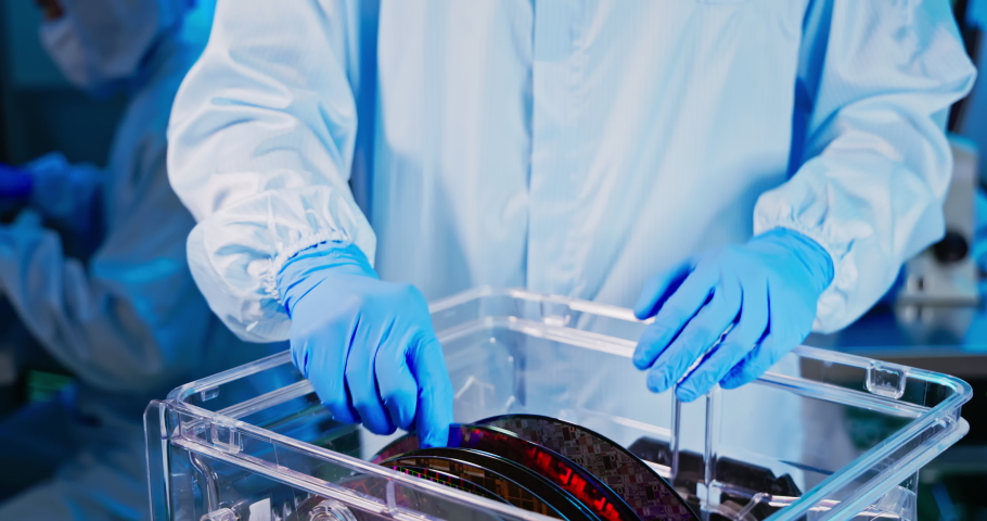 Close up of asian male technician in sterile coverall holds wafer with gloves that reflects many different colors and check it at semiconductor manufacturing plant | Shutterstock HD Video #1083484240