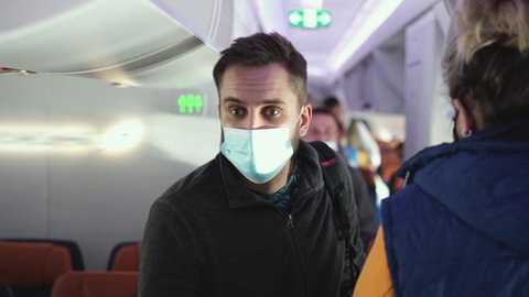 Male passenger in covid-19 face mask and backpack walking past other people to take his seat. Traveling, tourism during coronavirus pandemics. Travelers with maksed faces on plane board during flight
