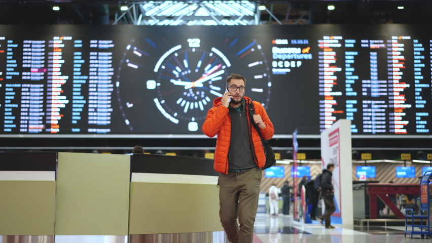 Travel or vacation passenger person in glasses wearing orange jacket with backpack talking on phone at airport terminal. People arrival on runway. Flight check technology, modern airport board sign. Royalty-Free Stock Footage #1083485299
