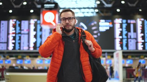 Travel or vacation passenger person in glasses wearing orange jacket with backpack talking on phone at airport terminal. People arrival on runway. Flight check technology, modern airport board sign.