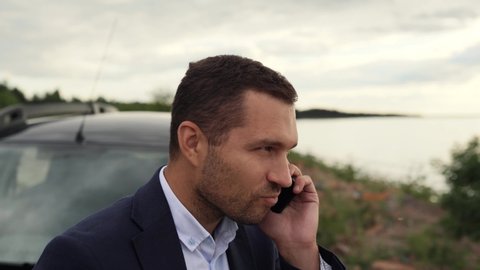 A business man stands near his car on the coast of a lake, sea or ocean, speaks on the phone, he is happy to new successes in his business