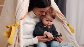 Close up of beautiful young mother and cute baby having video chat using smartphone mom sitting on cozy tent in living room enjoying mobile technology sharing motherhood lifestyle on social media