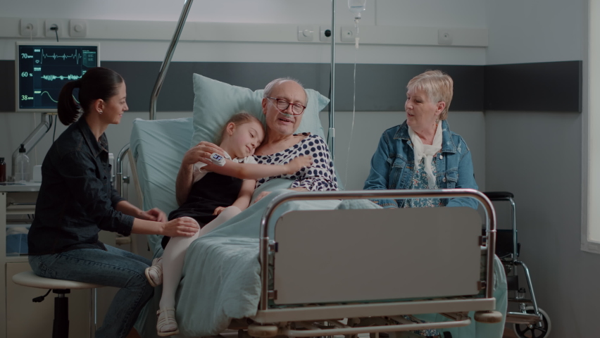 Niece hugging ill grandpa at visit in hospital ward bed. Little girl visiting senior man with sickness, giving support and helping with recovery. Retired patient enjoying assistance from visitors.