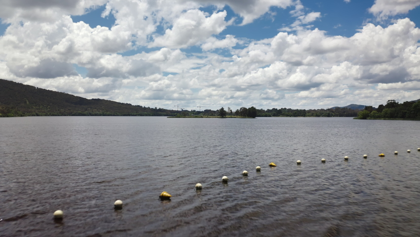Aerial drone shot flying low across the water towards a small island in the middle of a large lake, with hills and cloudy blue sky in the background. Located in the capital city of Canberra, Australia