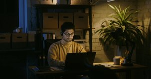 Online team game with microphone communication. Young handsome man with glasses dressed in casual clothes plays on a laptop online, talking with other players, under the light of a lamp