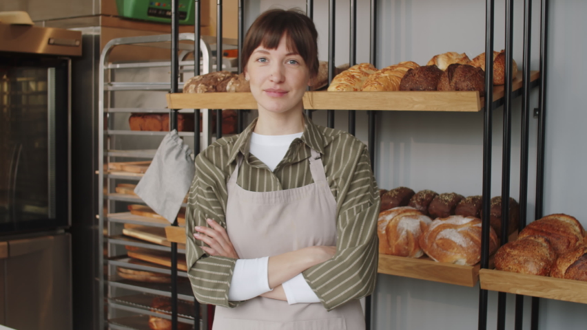 Waist up portrait shot of young beautiful woman in apron standing with arms crossed in bakery and looking at camera Royalty-Free Stock Footage #1083497944