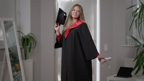 Smiling beautiful grad woman posing with rolled certificate and graduation cap indoors at home. Portrait of confident Caucasian graduate looking at camera smiling. Confidence and happiness