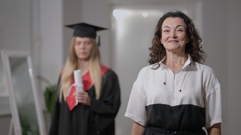 Excited Caucasian mother posing at home looking back at intelligent graduate daughter with rolled certificate. Portrait of happy glad proud woman looking at camera smiling. Support and intelligence