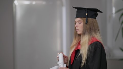 Side view cheerful Caucasian grad woman using rolled diploma as spyglass smiling. Happy smart young beautiful graduate having fun indoors at home on graduation day. Lifestyle and joy