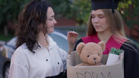 Worried graduate woman with dorm room box and mature mother supporting grad daughter standing outdoors talking in slow motion. Portrait of anxious Caucasian grad with parent