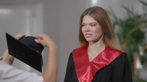 Portrait of smiling slim beautiful young woman looking at mother putting on graduation hat. Happy smart Caucasian daughter getting dressed for ceremony with parent indoors