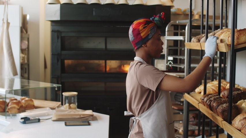 Young African American saleswoman in gloves putting fresh bread in paper bag, smiling and speaking with Caucasian female customer as she paying with smartphone in bakery Royalty-Free Stock Footage #1083502282