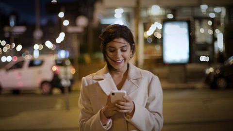 Close up Slow Motion of Attractive and Cheerful Businesswoman Using Smartphone at Night in Big City. Female Entrepreneur Walking and Texting in Cellphone on Windy Night. Traffic Lights. Frontal view.
