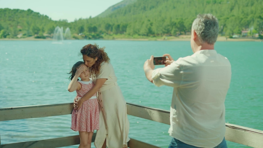 Cheerful mother and daughter posing for their father by the lake. A family spending time together in summer, hiking and enjoying nature and outdoor activities next to a beautiful mountain lake. 