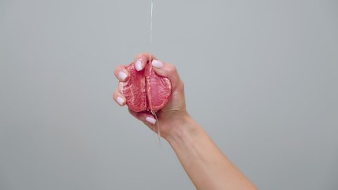 A woman's hand clutches half a grapefruit in her fist. Liquid is poured on the fruit. Close up. Gray background. 