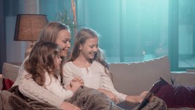 Beautiful woman sitting on couch with little girls and using modern laptop for video call. Caucasian mother and her two daughters smiling and waving hands during online conversation. 