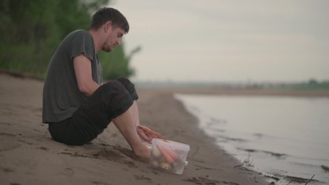 A man peels vegetables for cooking on a hike. Beautiful evening riverbank and sandy beach