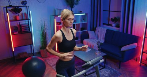 Attractive purposeful active fit slim young woman in training clothes wipping the sweat from forehead during running on treadmill at home in the evening