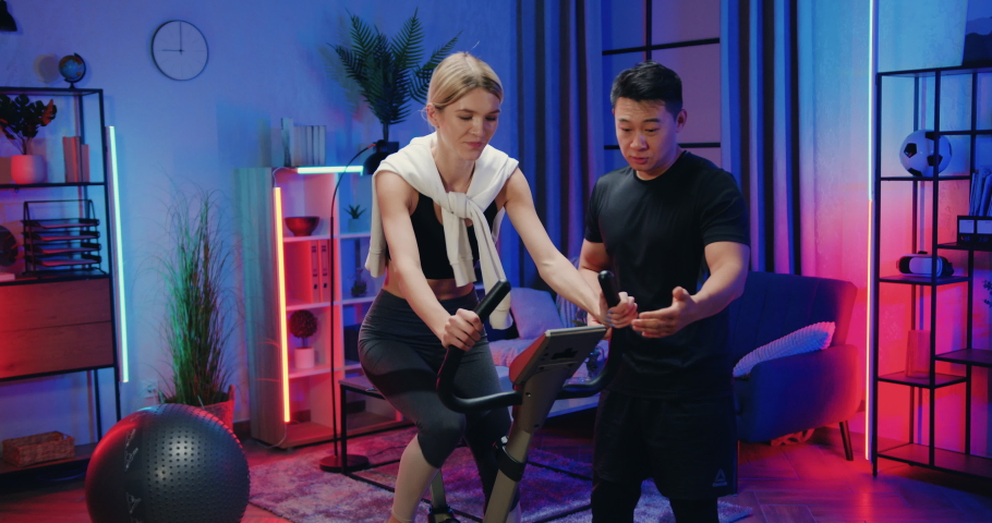 Good-looking confident athletic muscular asian guy setting the speed on stationary bike's control panel on which exercising his cute sporty active blond girlfriend at home in the evening