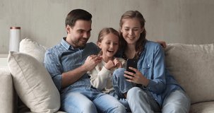 Happy friendly family of three mom dad and small daughter cuddle on soft couch at living room watch amusing video on modern cell. Bonding parents and little girl enjoy funny clips online using phone