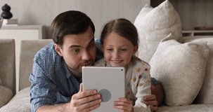 Single dad and preteen daughter lying close together on cosy couch hold digital tablet device engaged in educational activity. Interested father and girl using a digital tablet at home play game watch