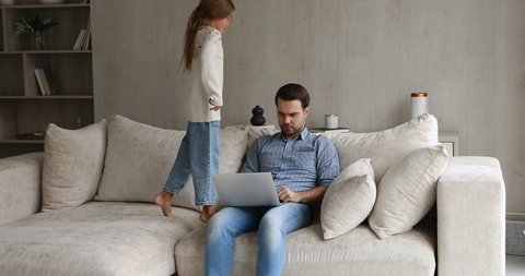 Serious dad work from home try to focus attention on pc screen read document online do business task while mischievous little girl jump on couch near. Small daughter behave bad disrupt job of father