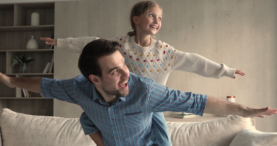 Playtime. Overjoyed young father carry excited little daughter on back pacing around living room spreading hands wide. Laughing small girl having fun riding on dad playing flying airplane game at home Royalty-Free Stock Footage #1083529954