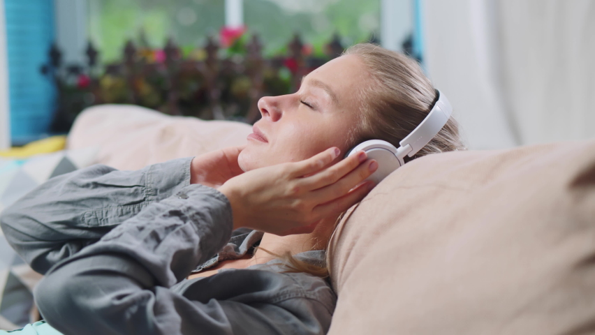 Young beautiful woman listening to music in headphones and relaxing on couch. Realtime. 
