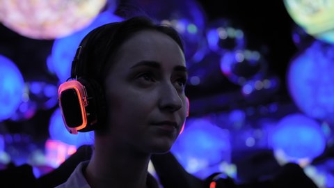 Woman wearing wireless black headphones and looking around in dark room of interactive exhibition or museum with colorful illumination - close up. Futuristic, immersive, entertainment concept