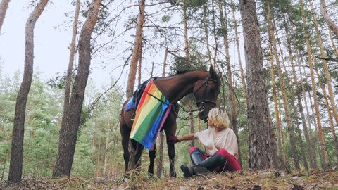 lgbt. rainbow flag. equestrian riding. beautiful rider girl, horsewoman is treating her horse with goodies, while sitting and resting during walking in the forest