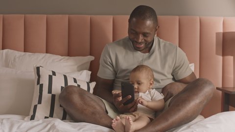 Full shot of young happy African man sitting on bed, showing smartphone to his adorable toddler son, smiling, then taking call