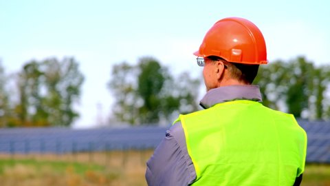 Solar station provides power in rural area. Builder in orange hardhat and yellow vest stands at construction site checking sun panels backside view