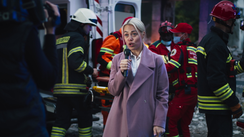 Blond woman in coat speaking into microphone near cameraman while emergency service workers saving survivors after earthquake