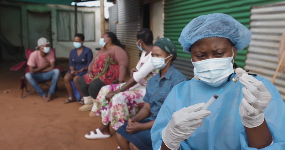 Black African female nurse pulling Covid-19 vaccine liquid from vial. African people line up for Covid-19 vaccine in a rural settlement. Royalty-Free Stock Footage #1083561679