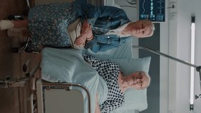 Vertical video: Portrait of senior man in hospital ward bed receiving assistance from wife, waiting on medical advice to cure sickness. Old patient and woman getting ready for consultation at clinic.