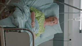 Vertical video: Portrait of old woman sitting in hospital ward bed to cure disease, being hospitalized at intensive care. Patient with nasal drip bag and heart rate monitor for healthcare and medicine
