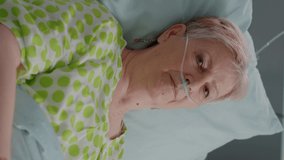 Vertical video: Close up of elder woman sitting in hospital ward bed with IV drip bag and nasal oxygen tube to cure sickness. Aged patient waiting to receive medical support and assistance against