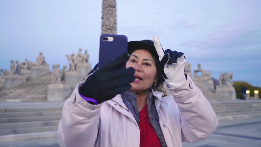 Asian woman standing and taking video call in Vigeland park, Norway. Beautiful day with beautiful sculptures and blu sky, Oslo