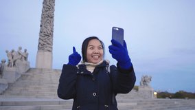 Happy Asian woman standing and taking video call in Vigeland park, Norway. Beautiful day with beautiful sculptures and blu sky, Oslo