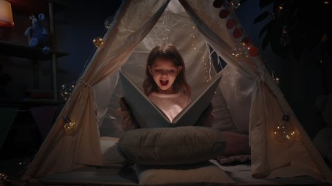 Pretty little kid in cute makeshift hut having fun in her room in evening. Beautiful child sitting on floor while reading book with flashlight. Concept of leisure and careless childhood