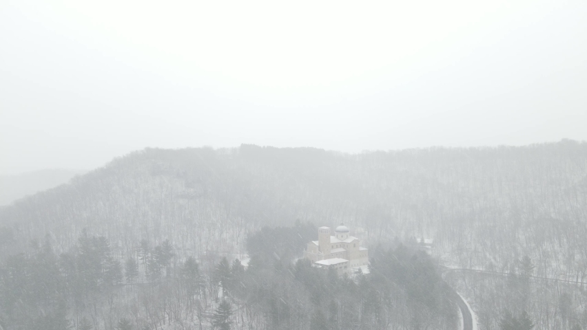 Drone view of snow falling over forest and mountain in Wisconsin. Road leading up hill. Church through the trees. White sky from snow falling. Groups of pine trees.