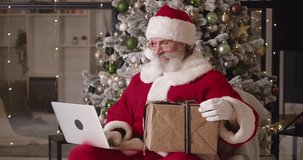 Cheerful Santa Claus sitting on an armchair against a Christmas tree background in his residence, chatting online during video call, showing thumb up and a Christmas gift box to his interlocutor