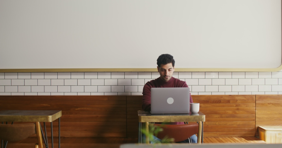 Young dark-haired man with thick stubble on his face, dressed in casual clothes, uses a laptop while sitting at a small table in a cafe, working remotely Royalty-Free Stock Footage #1083604819