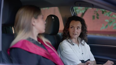 Portrait of confident mature woman sitting in car talking to blurred graduate on passenger seat. Happy proud Caucasian mother giving advice to daughter in slow motion